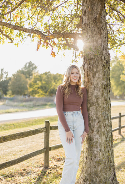 high school senior female, leaning against a tree, smiling at the camera, wearing jeans and long sleeve shirt, sun is setting behind her