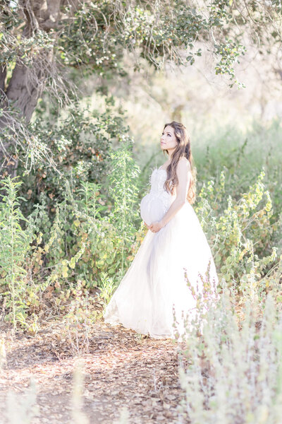 A pregnant woman in a long white dress standing in a forest