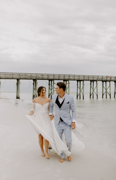 newlyweds walk on beach in front of pier