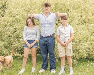 Two brothers and a sister standing in the grass in front of a bush.  Oldest is wearing a light blue button up shirt, blue jeans, white shoes, closed smiling at the camera, and has his arms on his siblings heads giving thumbs up.  Younger brother is wearing a light tan button up shirt, khaki shirt, white shoes, and smiling at the camera.  Sister is wearing a 3 quarter sleeve light blue shirt, jean shorts, white shoes, dark hair, smiling at the camera.  