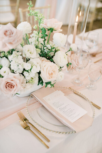 A table setting curated by a Banff wedding planner features a floral centerpiece with white and blush pink roses, a menu card on each plate, gold-colored cutlery, and elegant glasses. A lit candle softly illuminates the scene, showcasing the attention to detail that wedding planning services provide.