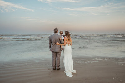 Bride and groom hold their young son as they gaze out to the ocean and watch the sun set.