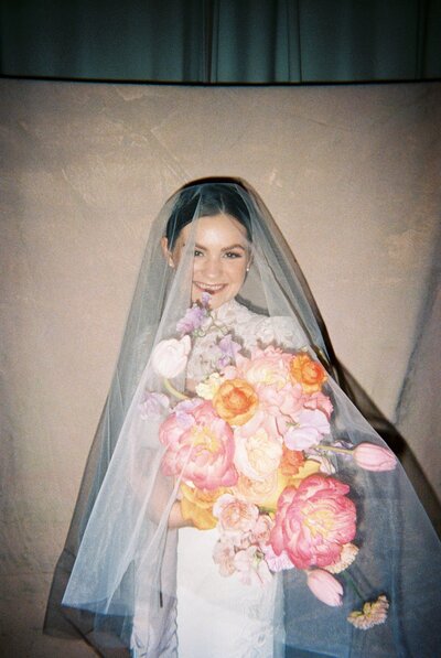 Flash photo of bride with veil over her face holding up large pink and orange bouquet
