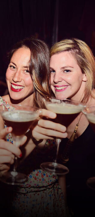 Close-up of two women smiling and raising espresso martinis toward the camera during a lively celebration.