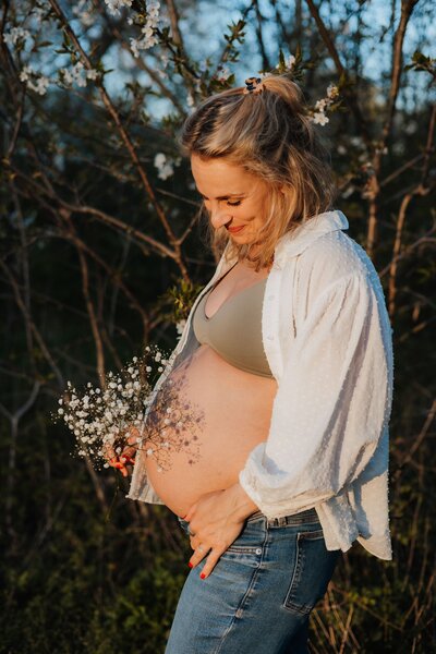 Golden Hour Shooting von Mutter in weißem Hemd, sie hält Blumen über ihren Babybauch