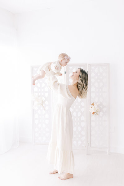 mother in cream colored dress holding one year old daughter in the air in white studio in scottsdale arizona