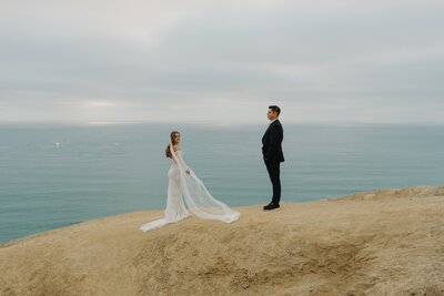 bride and groom standing apart on san diego sand cliff with ocean 