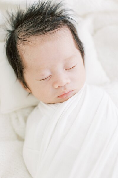 Newborn asian baby with a head full of hair swaddled in white sleeping in moses basket for newborn photos