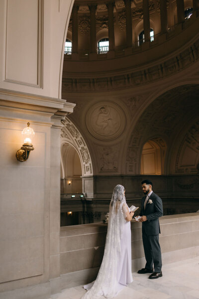 A bride and groom hold hands in San Francisco City Hall with ornate architecture, under a dome and beside a classical wall mural and elegant light fixtures