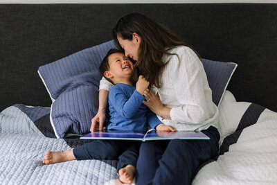 mom and son sitting on bed holding a book with their faces close, snuggling, and looking into each other's eyes
