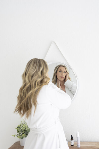 Woman in a white robe looking in the mirror, preparing for her morning skincare routine—representing The Budget Dermatologist’s approach to simple, real skincare.