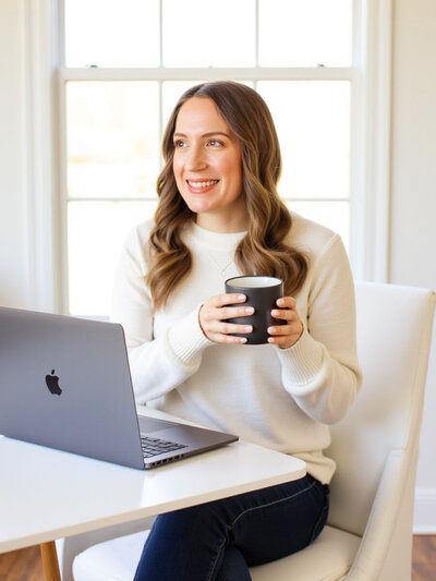Stacey from Xanthe Bookkeeping smiling in a white sweater, sitting at a table with a MacBook Pro and holding a coffee cup.