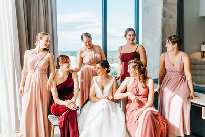 Six attendants in flattering shades of pink and a bride in a white dress sit closely together at the Watermark Hotel in Tysons Corner, Virginia Wedding Makeup