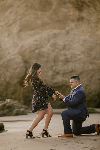Man in suit on bended knee, proposing to surprised and happy girlfriend, on beautiful beach setting