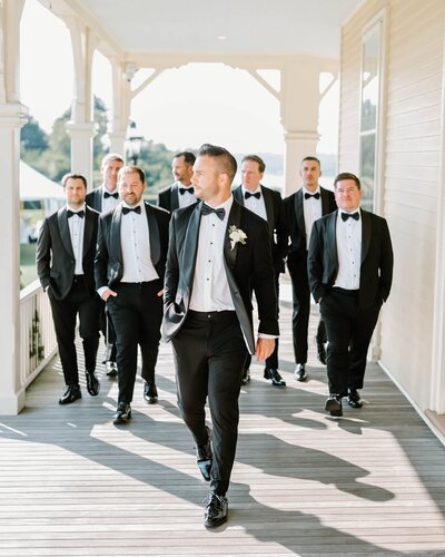 Newport Wedding Photographer | A groom in a tuxedo walks confidently on a sunlit porch, followed by seven groomsmen in matching black suits.
