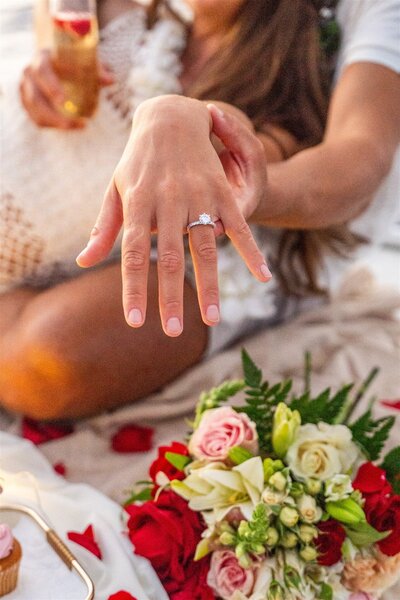 Close-up of a woman showing her engagement ring while relaxing on a sailboat in Hawaii, with rose petals and a bouquet beside her during the proposal celebration.