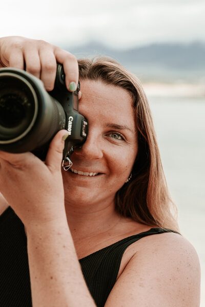 Sierra Bertini looking through her camera while taking a photo half of her face is covered by her camera on a beach in Maui