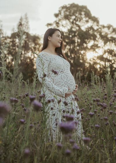 Pregnant women in a white long crochet dress that shows off the beautiful baby bump that she's cradling at her outdoor maternity photo session.
