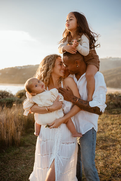 A family of four stands outdoors at Umina Beach near a body of water, with a woman showcasing her radiant maternity glow while holding a baby and a man happily carrying a young girl on his shoulders. Everyone appears happy and relaxed.