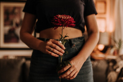A woman holds a red flower in front of her torso.