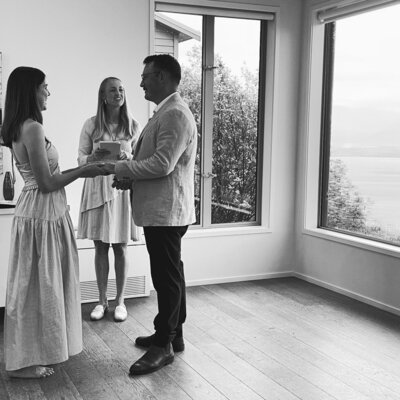 A black-and-white photo of a couple standing indoors during an intimate wedding ceremony, holding hands and exchanging vows in front of a large window with a scenic view, while an officiant stands between them.
