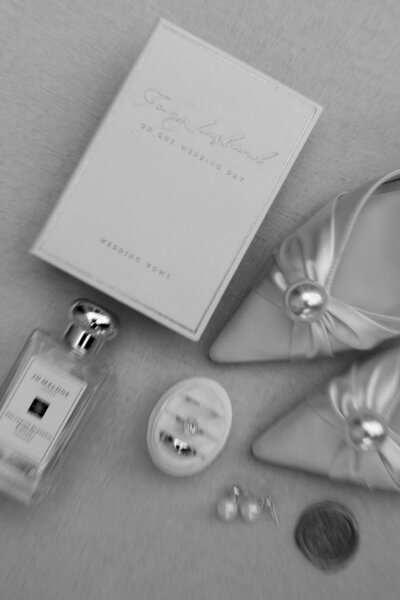 Flat-lay of elegant wedding details including vow book, pearl heels, jewelry, and perfume — captured by a destination elopement photographer.