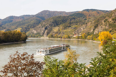 A white river cruise ship travels along a wide, calm river surrounded by autumn-colored trees and rugged hillsides under a light, hazy sky.