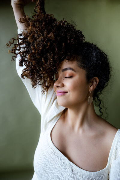 Studio portrait of a woman with curly hair, smiling softly while posing against a muted green background.