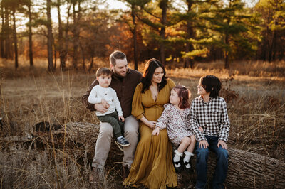 family of five posing for a fall image outside on a log with tall trees and the sun setting behind them. They are all looking towards each other while Paula Goforth takes their fall family pictures in Decatur 
