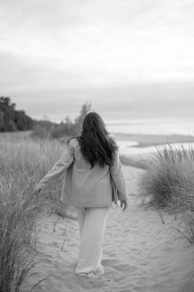 A woman in an oversized blazer and trousers walking through Lake Michigan dunes, grazing her hands across the dune grass.