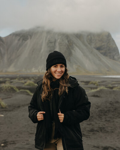 Destination elopement photographer smiling on a black sand beach with misty mountains in the background, wearing a black jacket and beanie in a moody, adventurous landscape.
