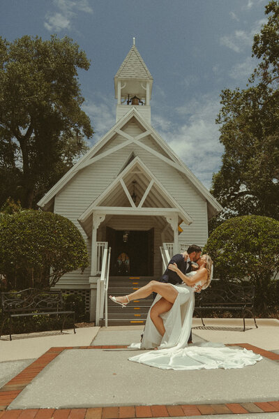 Bride and groom sharing a cinematic dip kiss in front of a charming white chapel, bride in a flowing gown and groom in a navy suit on a sunny Texas day.