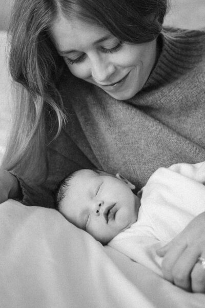 Mother gazes down at her sleeping newborn with a soft smile during a quiet in-home newborn session in Munich.