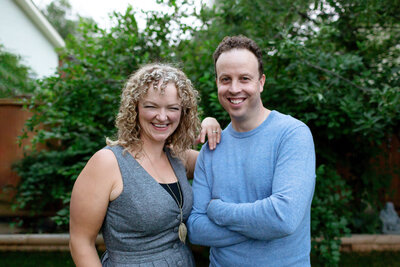 Calgary brand photography team shot of Switchback Creative, husband and wife, who are standing beside each other and smiling at the camera. They are wearing blue and grey and there are green leaves behind them in their backyard.