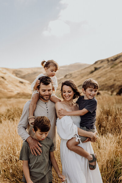 A mother and father holding their three kids, the daughter is riding on the dads shoulders and the younger brother is being carried by the mother. They're standing in long grass and smiling at one another while lucy jane photographs them in christchurch