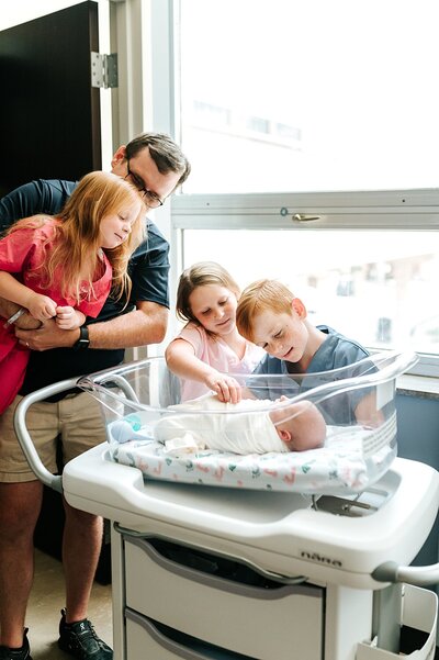 3 siblings and dad check out new baby who is lying in bassinet in hospital room