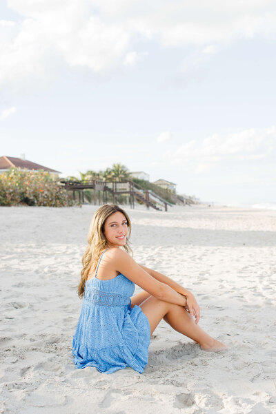 Senior wearing a beautiful blue dress and sitting in the sand smiling at sunset