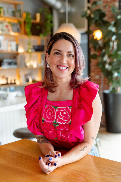 Vanessa Vancour, a Latinx woman with shoulder length hair,stands inside a restaurant wearing a pink shirt, leaning on a table and smiles. 