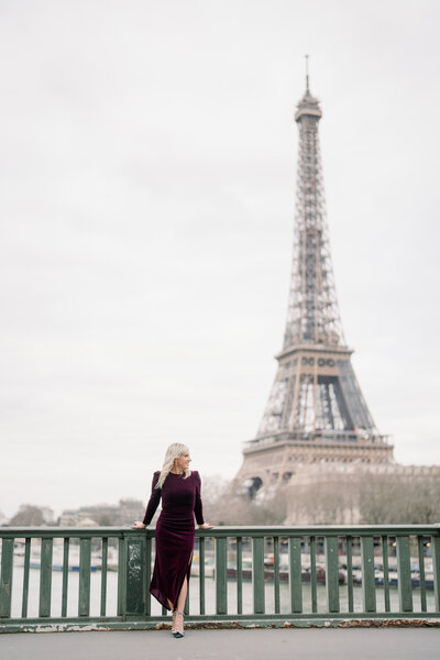 a blonde woman in a burgundy velvet dress looking off into the distance while holding the rails on the bir hakeim bridge with the eiffel tower behind her