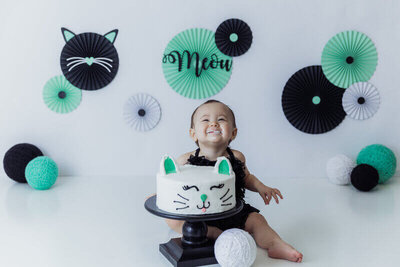 A smiling baby with frosting on their face sits behind a cat-themed cake with "Meow" decorations in the background.