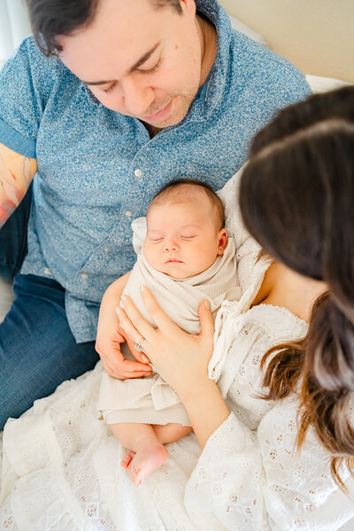 Austin Lifestyle Newborn Photographer | A couple sits on their bed holding their newborn baby, swaddled in a beige blanket.