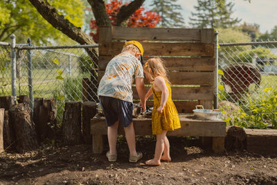 young brother and sister play in mud kitchen facing away from the camera