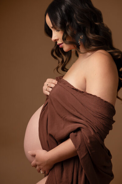 A maternity portrait featuring a side profile of a pregnant individual wrapped in a flowing, earthy-brown fabric.