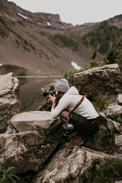 Janelle Renee, Washington elopement photographer, crouched down  on boulders aiming camera to take a picture