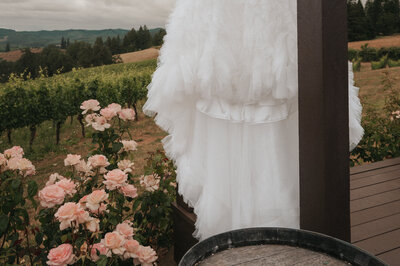 Elegant white wedding dress hanging beside blooming pink roses on a vineyard deck, with rolling hills of the Oregon wine country in the background, captured by a Portland wedding photographer specializing in Pacific Northwest vineyard weddings