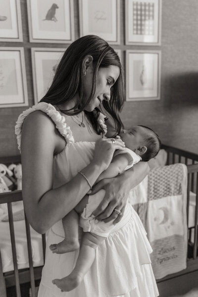 Black and white photo of mom standing beside crib while holding baby, eye contact between them.