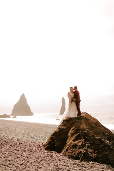 Elopement on the beaches of Iceland, bride and groom kissing while standing on a rock, with rock formations in the water behind them
