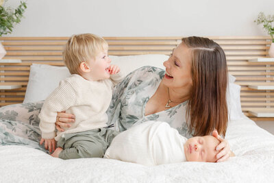 Mom laying down on bed with newborn baby in front of her and toddler son. Baby is sleeping soundly and Mom and Toddler are looking at each other and laughing, They are dressed in soft sage greens and creams and whites.