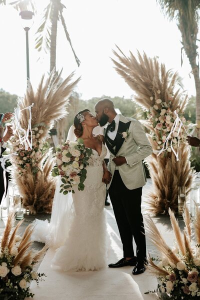 A bride and groom kiss under a palm tree during their wedding ceremony.