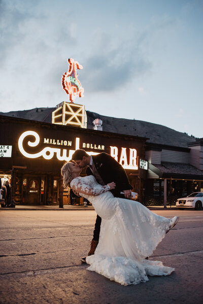 cowboy car in downtown jackson hole with groom dipping his bride and passionately kissing  his bride in the road in front of the bar captured by jackson hole photographers
